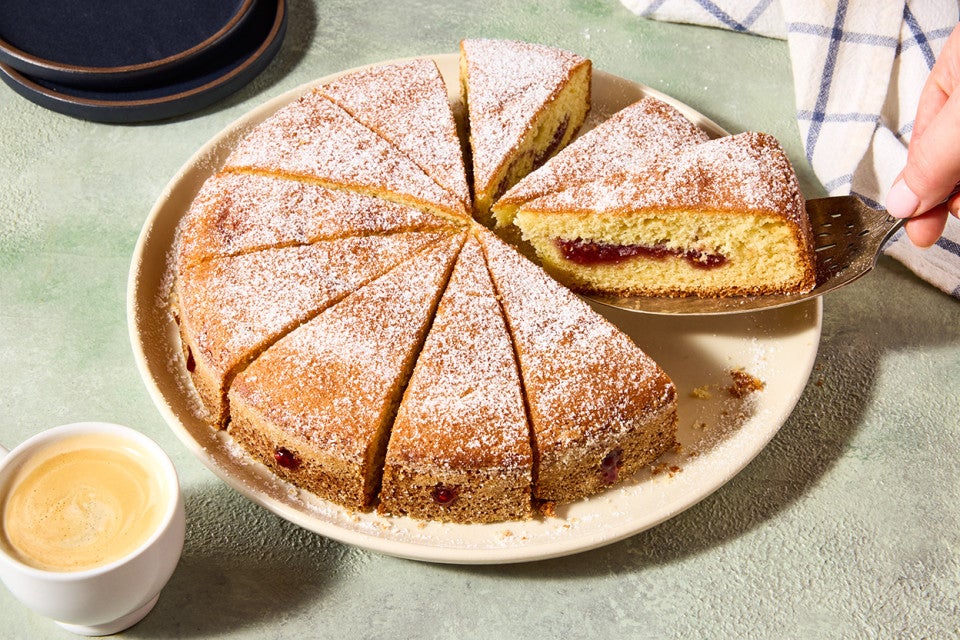 A hand removing a slice of Jelly Doughnut Cake from a plate. You can see the inside of the slice with a golden interior and lovely jelly filling.