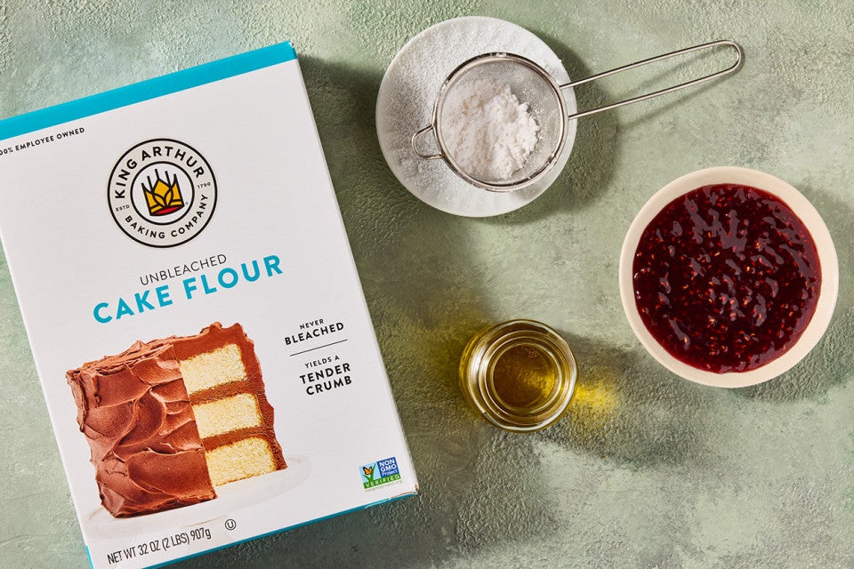 Cake flour, dry ingredients in a sifter, a bowl of jelly filling, and a cup of oil arranged on a counter.