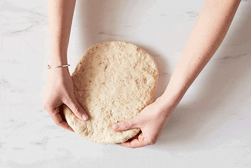 A baker shaping dough into a sandwich loaf by patting the dough out into a rectangle first, then pulling the top corners down to the center, tucking and rolling the dough down into a log.