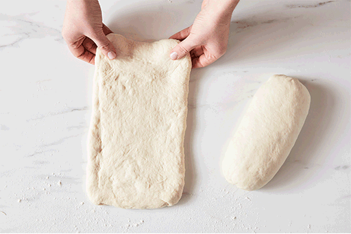 A baker folding dough into a loaf shape by first folding the dough in thirds (like a letter) and then using the heel of their hand to seal the seam.
