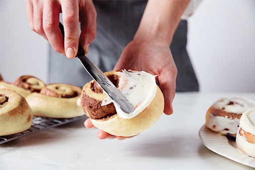 A baker holding one roll and using a small offset spatula to spread icing on top. 