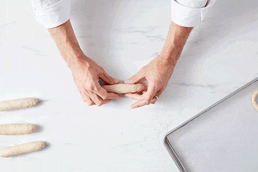 A baker rolling a log of pretzel dough into a long rope with tapered ends. 