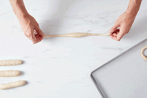 A baker shaping a pretzel by bending the rope of dough into a "U" shape, and twisting the ends together in the middle twice, then pressing the ends of the rope into the rounded part of the "U" made with the dough. 