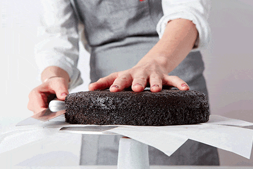 A baker using a serrated knife to split a cake into two thin layers.