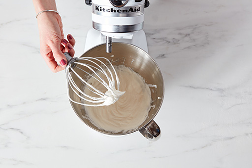 A baker holding up a whisk attachment for a stand mixer, showing the stiffness of the whipped cream. 