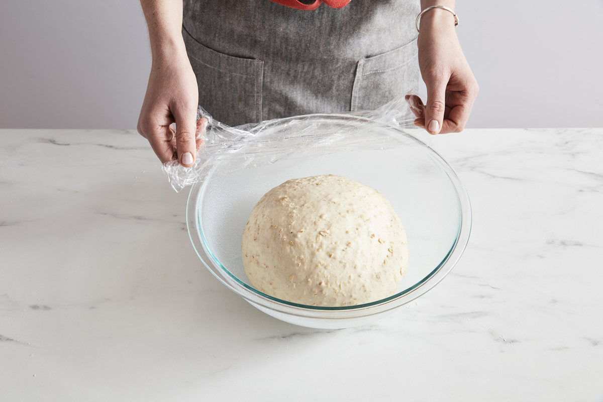Bread dough in a bowl being covered with plastic wrap.