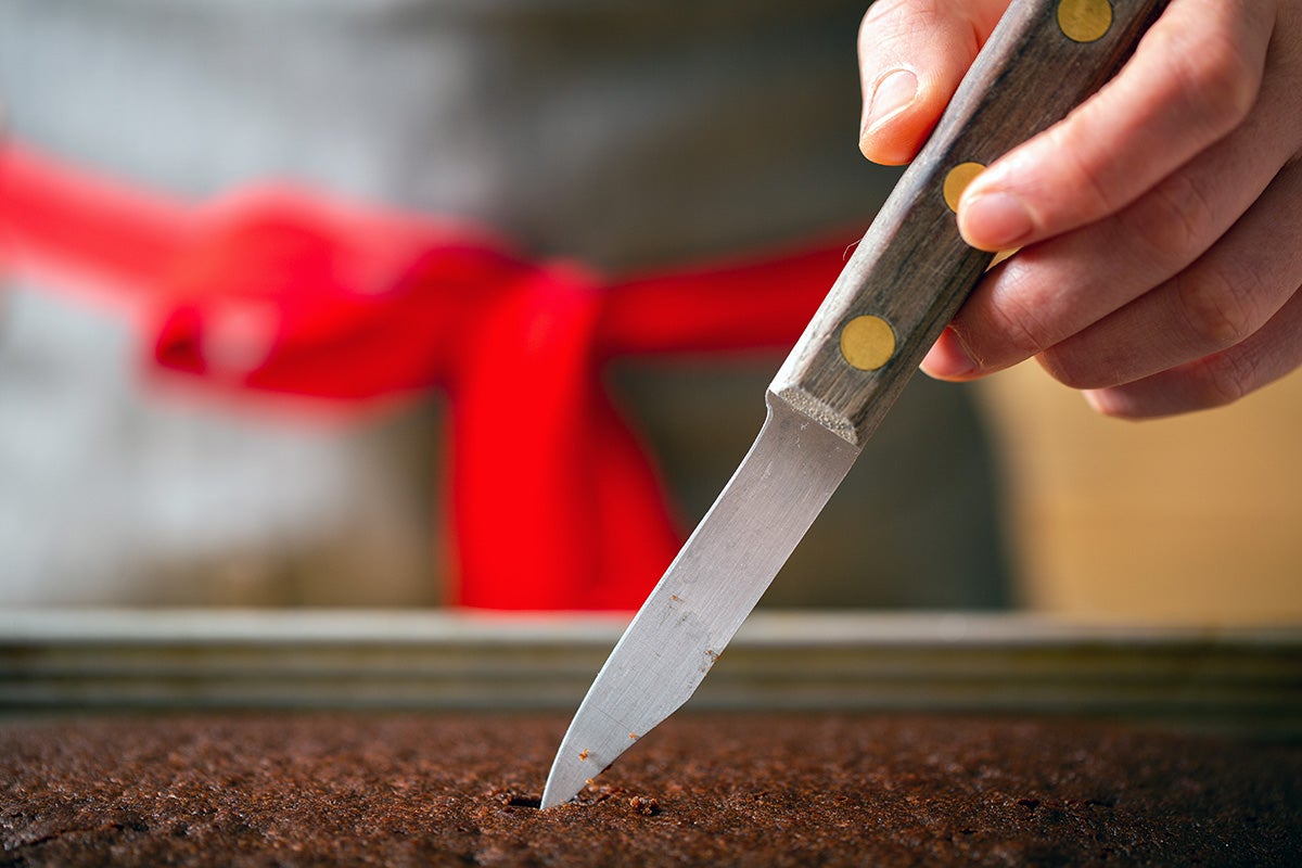 A baker inserting a paring knife into a chocolate cake to see if it's done baking