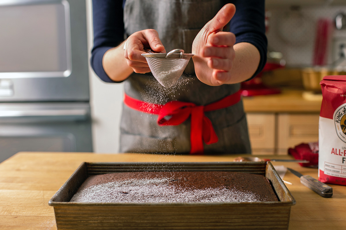 A baker's hands dusting confectioner's sugar over a baked chocolate cake