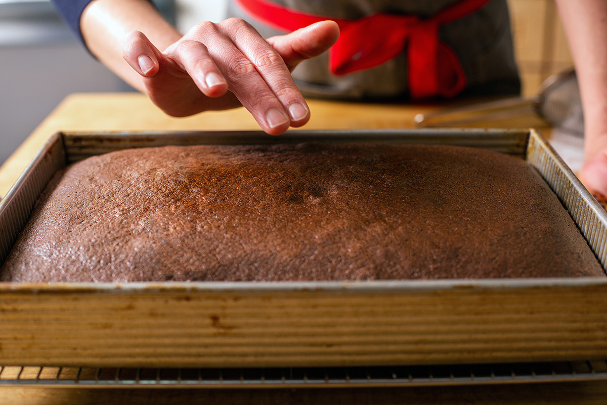 A baker pressing on the center of a chocolate cake that's not yet done baking