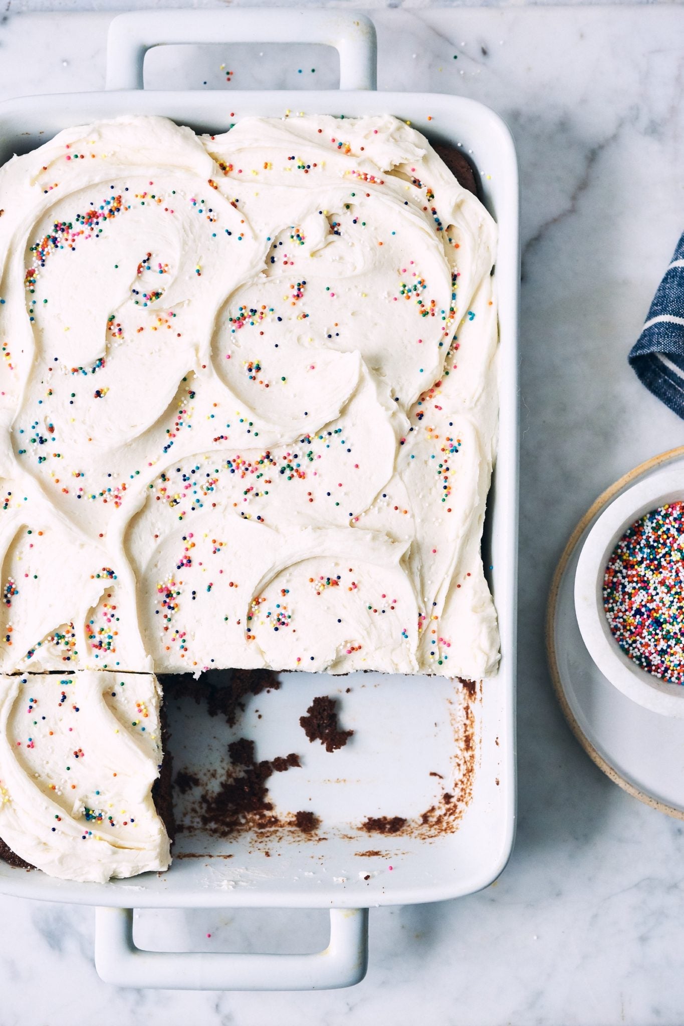 A chocolate sheet cake topped with vanilla frosting and rainbow sprinkles, a few slices removed