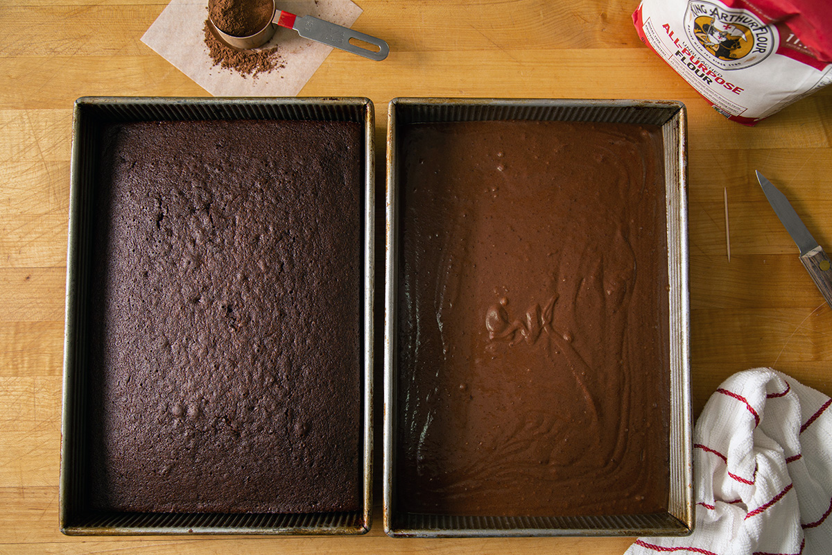 A fully baked chocolate cake next to an unbaked chocolate cake on a wooden table with flour and cocoa powder