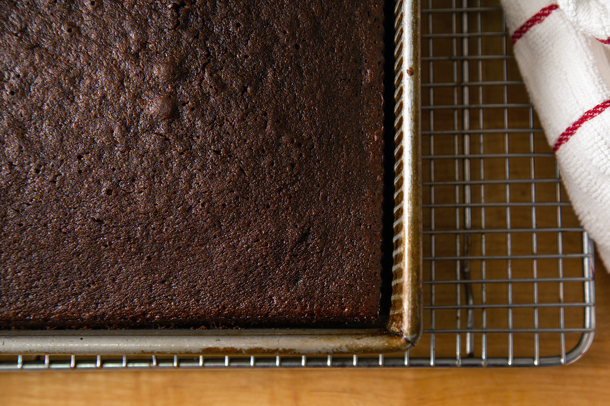 A pan of chocolate cake on a cooling rack with a small gap between the sides of the pan and the cake