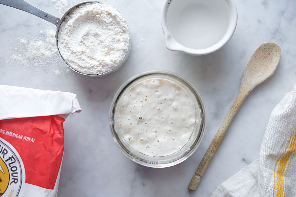 Sourdough starter on a counter with a cup of flour and a wooden spoon.