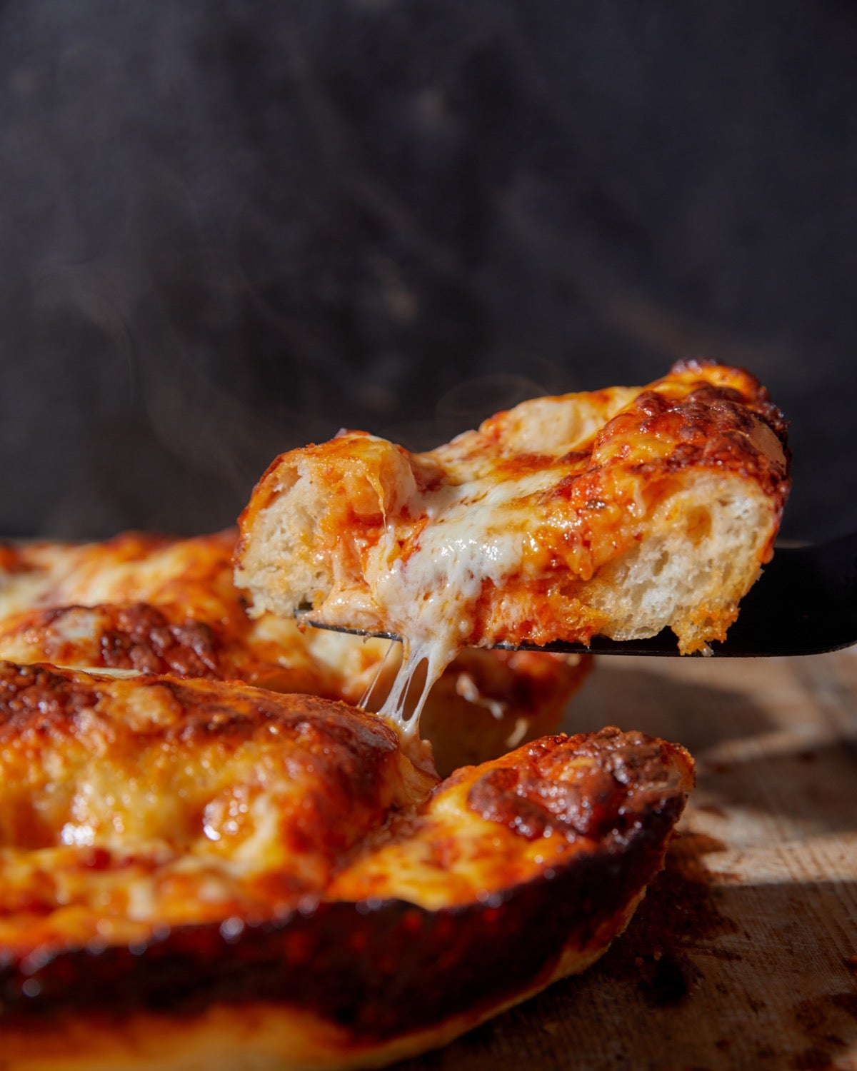 Thick-crust pizza on a cutting board, cut wedge being lifted out, cheese oozing.