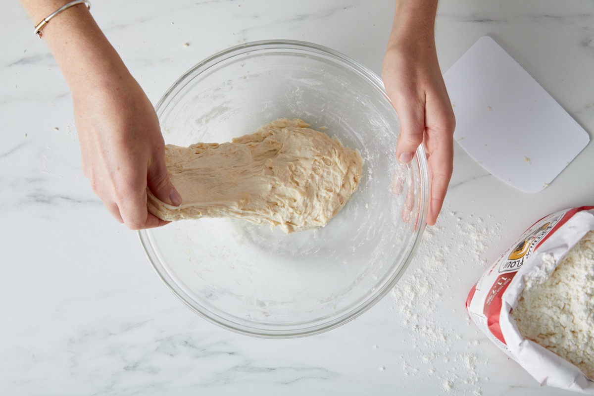 Dough in a glass bowl, a hand lifting one edge preparatory to folding it into the center.