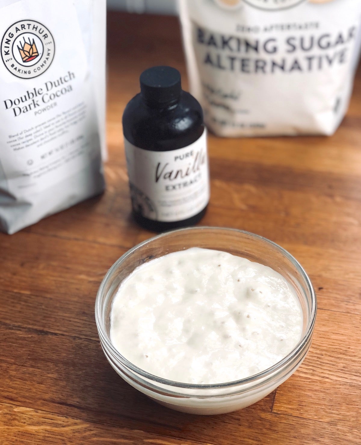 Bowl of sourdough starter with cocoa powder, vanilla, and baking sugar alternative in the background.