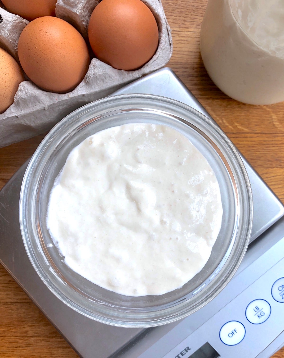 Bowl of sourdough starter on a scale, eggs in the background.