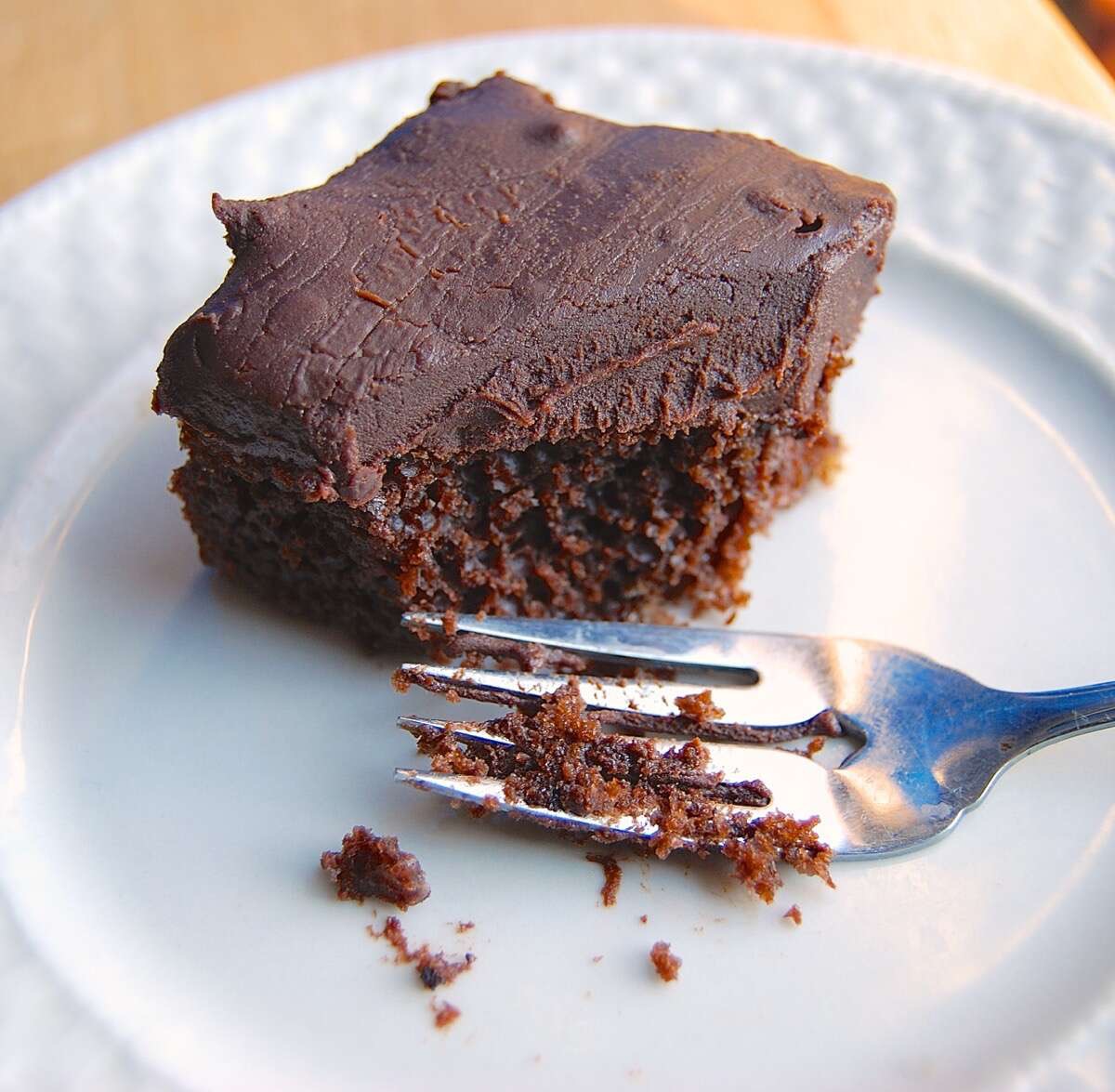 Square of chocolate cake with fudge frosting on a plate with a fork.