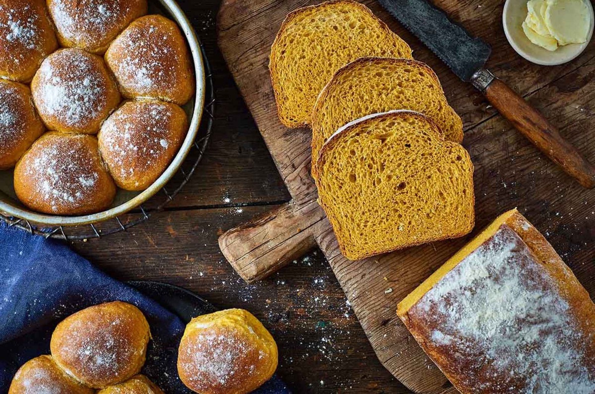 Loaf of pumpkin yeast bread, sliced, pan of rolls alongside.