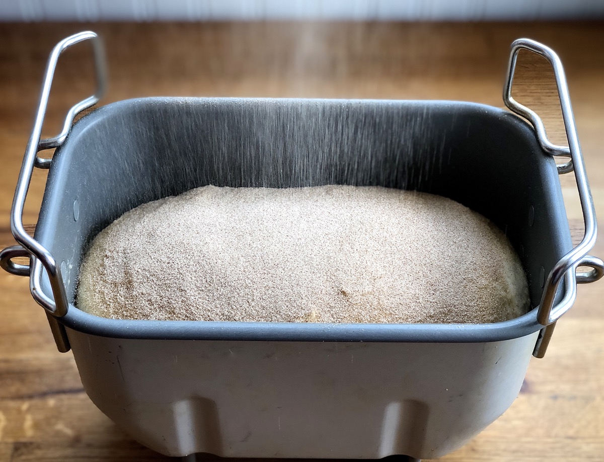Risen bread in bread machine being topped with cinnamon sugar prior to baking
