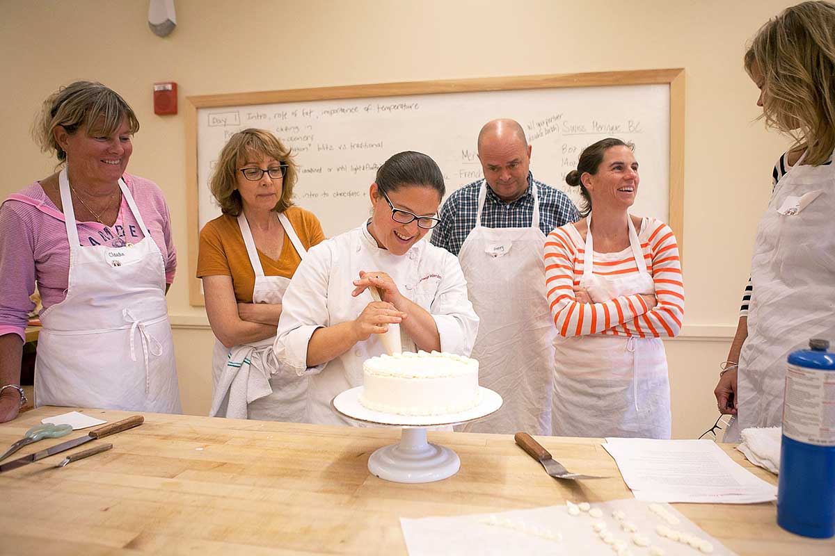 Baking school instructor frosting a layer cake, with students observing around her