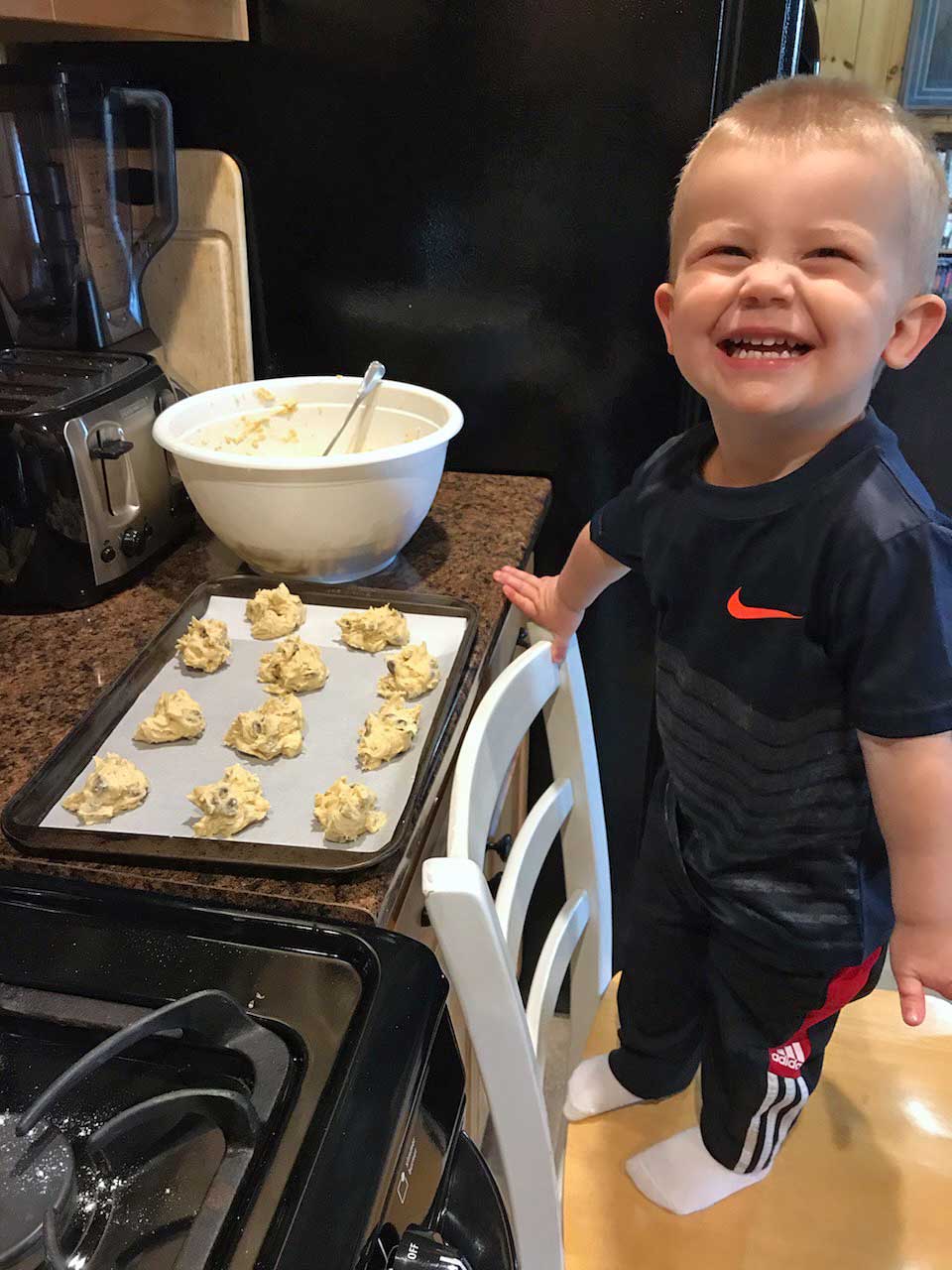 A small baker standing on a stool over a bowl of cookie dough and smiling
