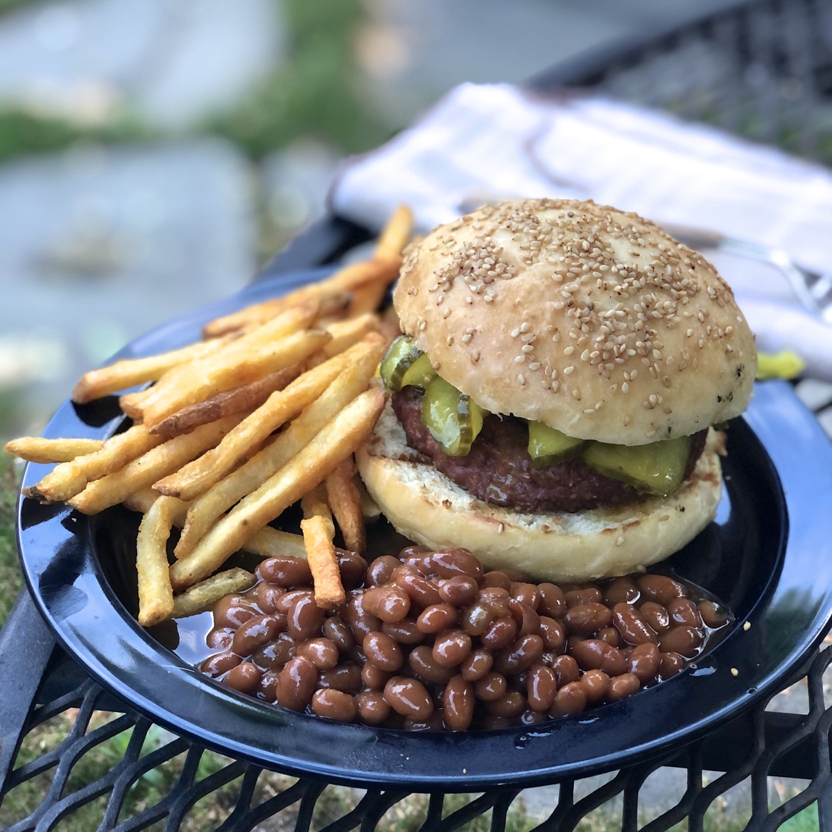 A burger in a bun with pickles, baked beans and fries on the side, on a blue plate on a picnic table.