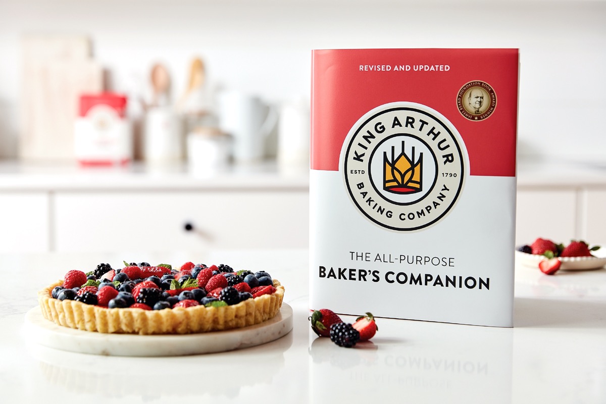 Baker's Companion cookbook sitting on a white table alongside a berry cream taert.