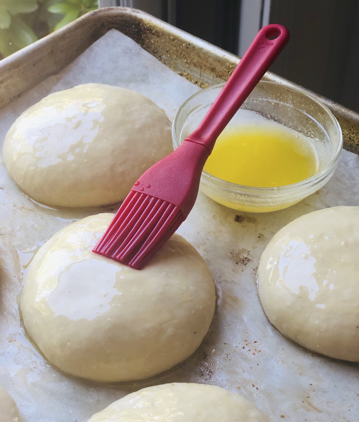 Risen  buns being brushed with melted butter using a red silicone pastry brush.