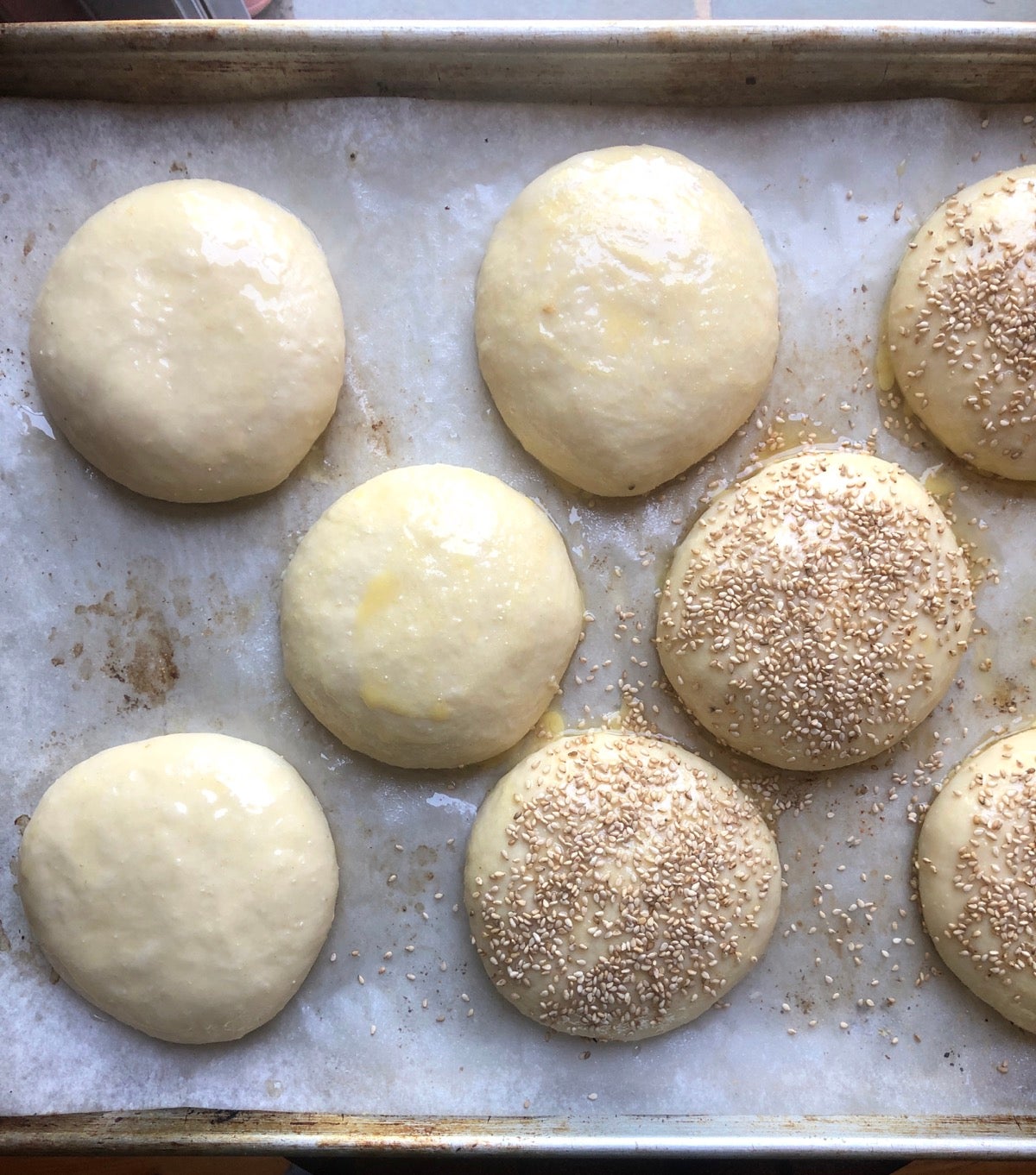 Risen buns on a baking sheet, brushed with butter, some sprinkled with sesame seeds, ready to bake.