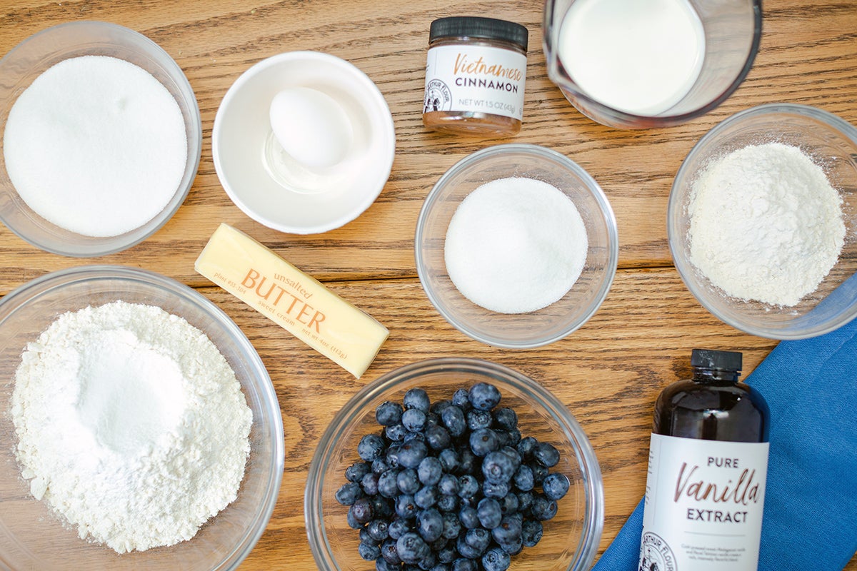 Bowls containing ingredients needed to make Blueberry Buckle Coffeecake: flour, sugar, butter, an egg, blueberries, cinnamon