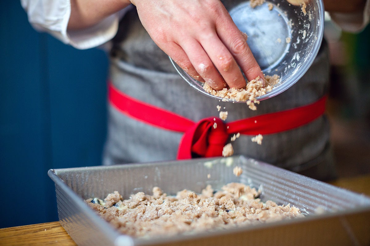 A baker sprinkling cinnamon streusel topping onto Blueberry Buckle Coffeecake batter