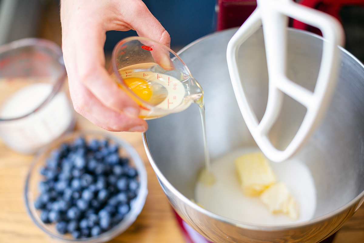 A baker pouring an egg into a mixing bowl with the Blueberry Buckle Coffeecake batter
