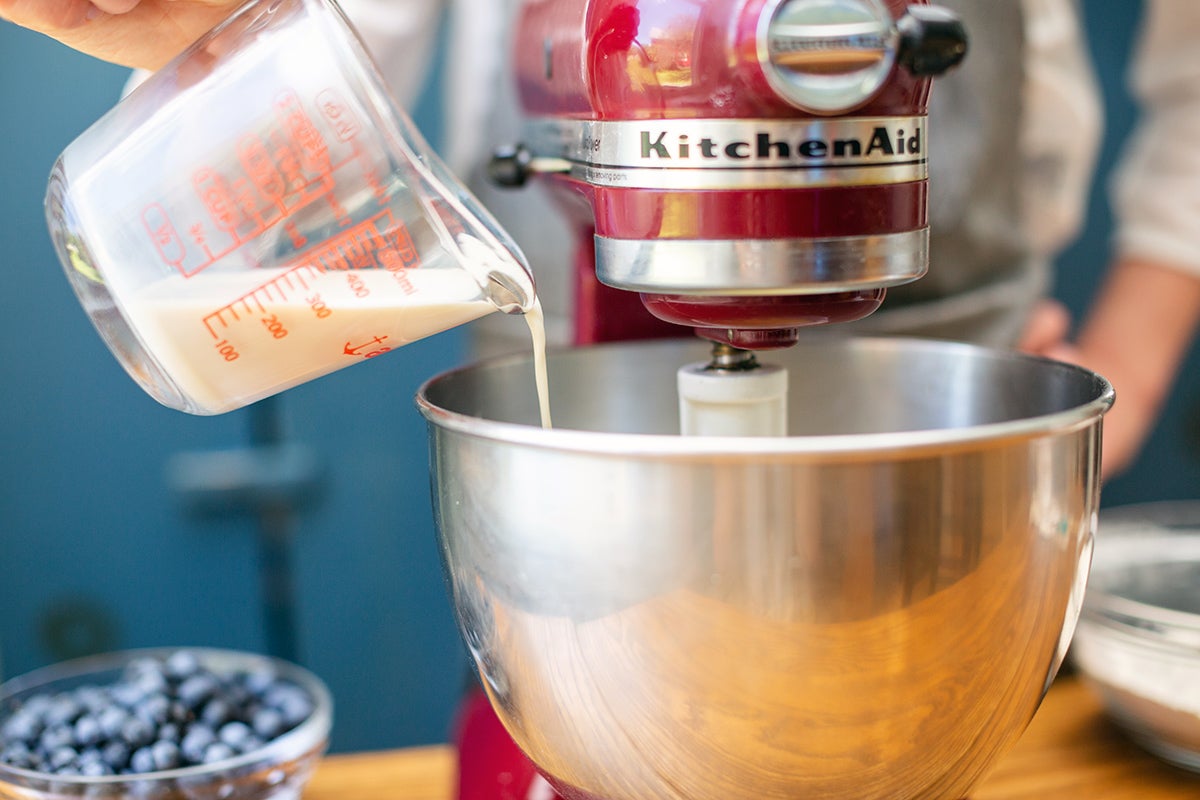 A baker pouring milk into a mixing bowl with Blueberry Buckle Coffeecake batter