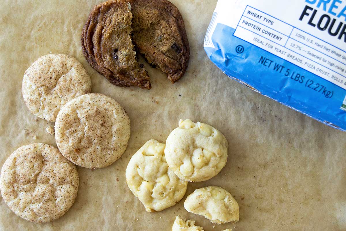 Buttery snickerdoodles, chocolate chip cookies, and white chocolate cookie drops on parchment next to a bag of bread flour