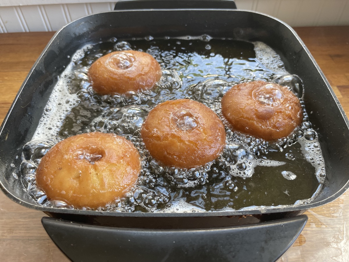 Cake-style doughnuts frying in an electric skillet of hot oil.