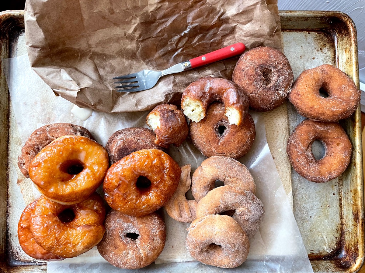 A baking sheet lined with paper towels and a paper bag, piled with just-fried doughnuts, both yeast-raised and cake-style.