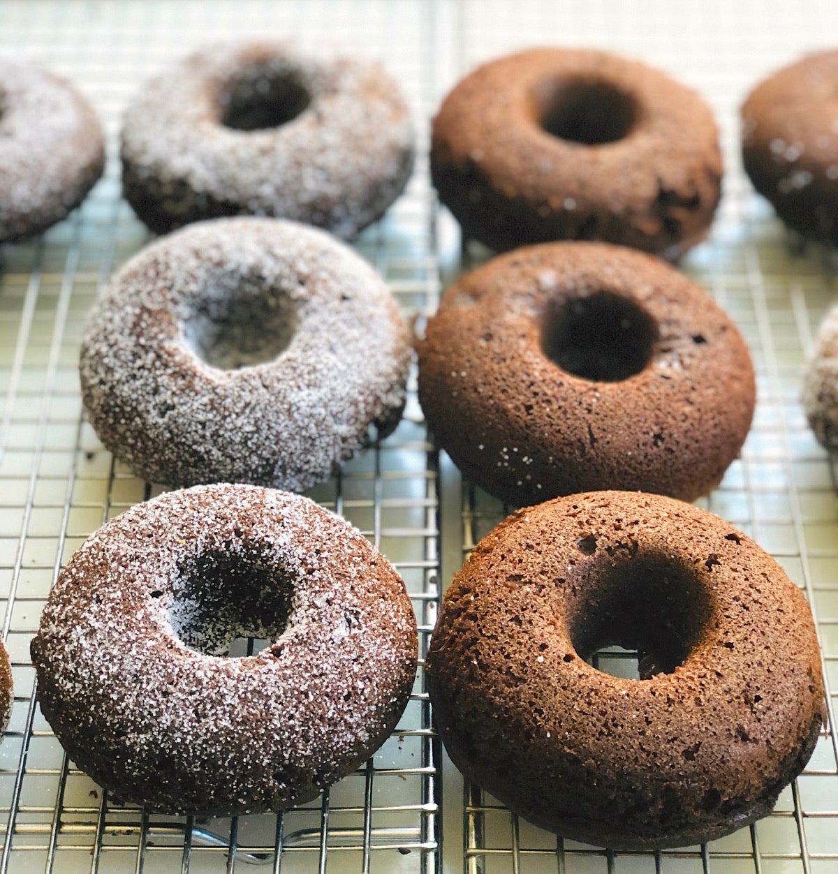 Chocolate doughnut son a cooling rack, half sugared with granulated sugar, half plain.