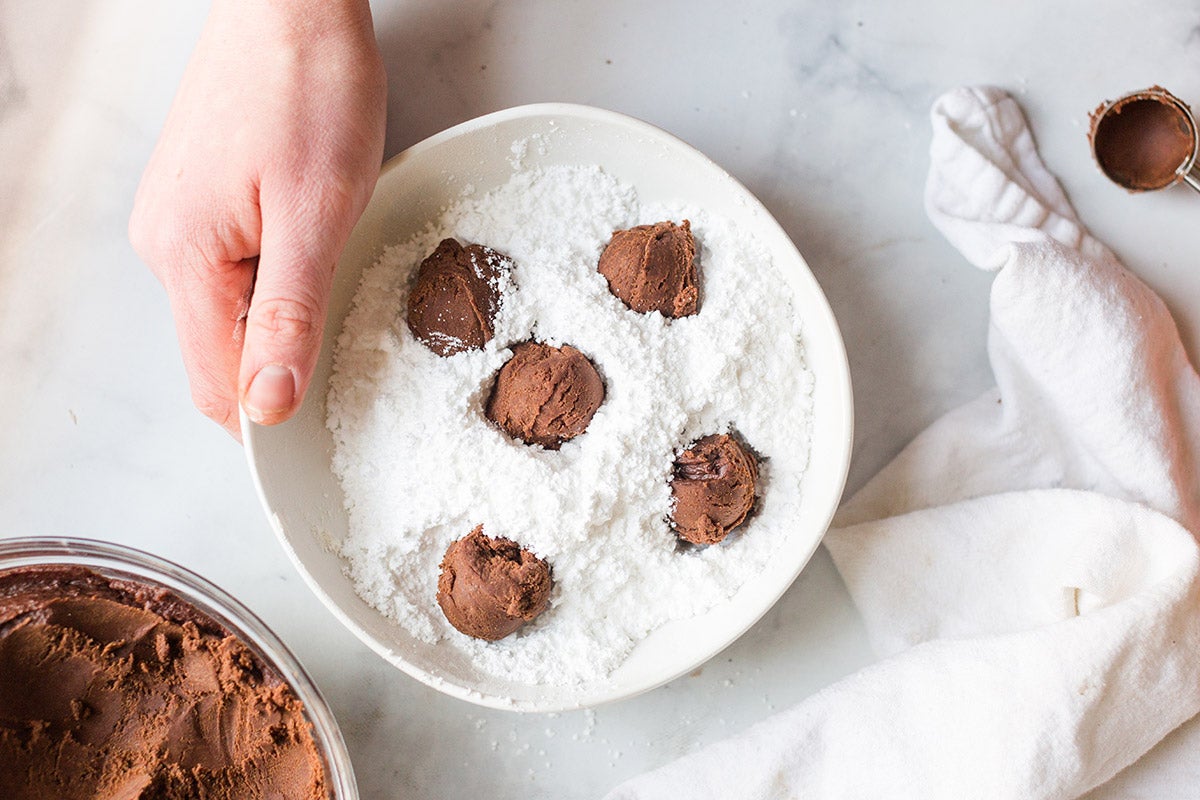 A baker holding a small bowl of confectioners' sugar with five balls of Chocolate Crinkle dough, ready to be tossed together