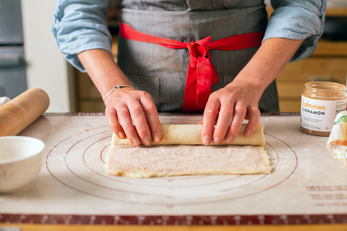 A baker rolling up pie dough to encase a cinnamon-sugar filling
