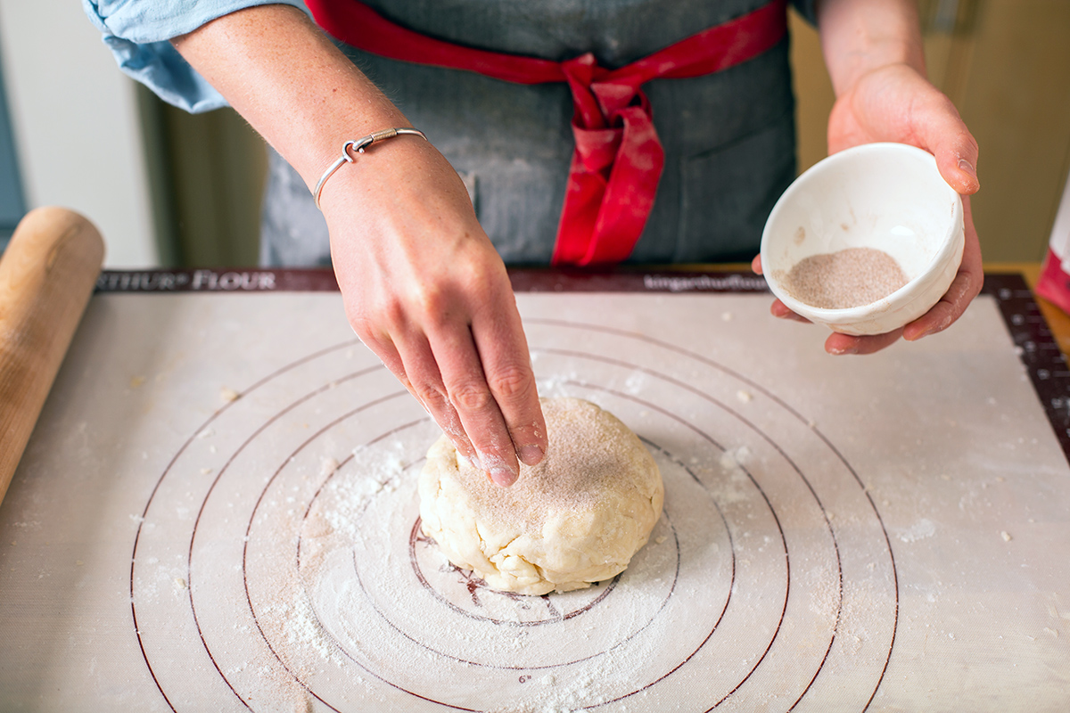 A baker sprinkling pie dough with cinnamon-sugar before rolling it out