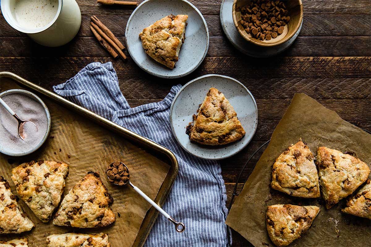 A selection of cinnamon scones on a kitchen table, surrounded by mugs of frothy coffee