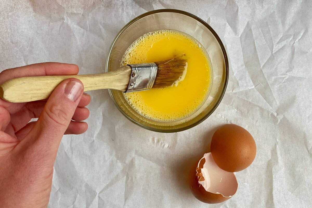 A baker dipping a pastry brush into a small bowl of egg wash