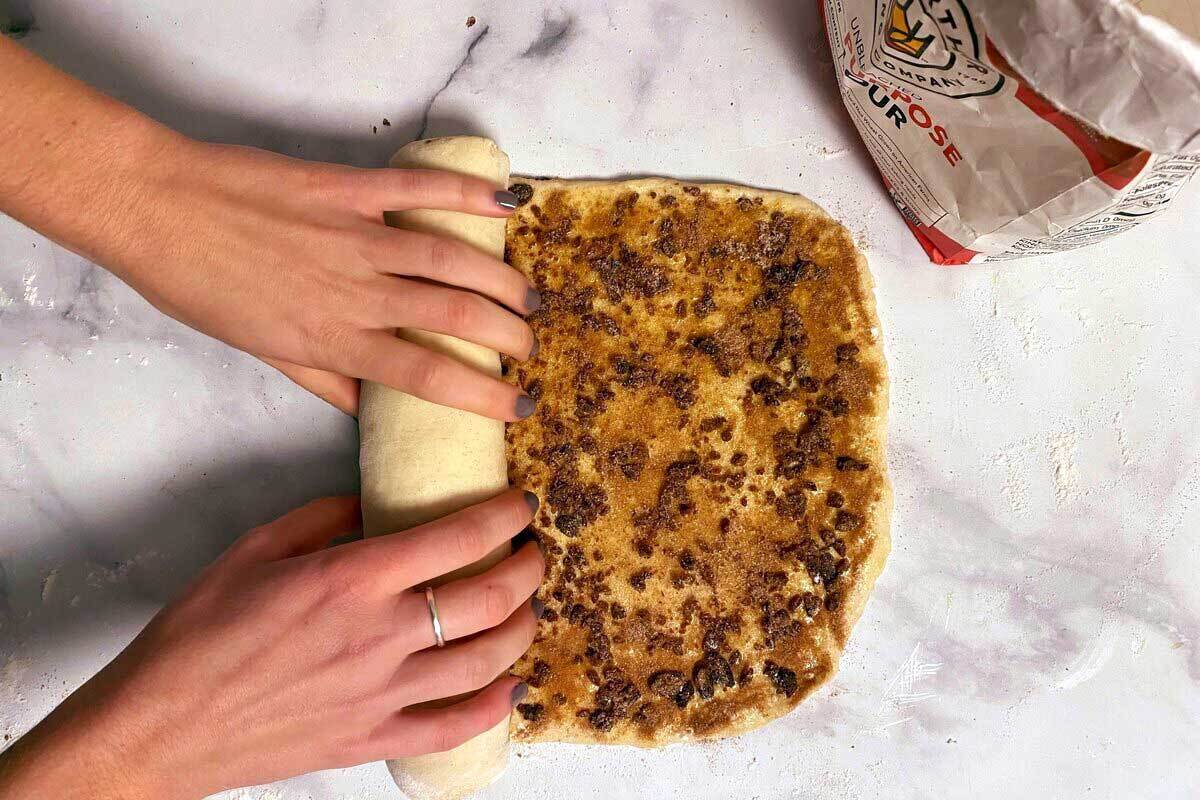 A baker rolling up a log of cinnamon swirl bread dough