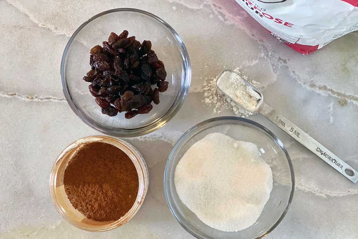A small bowl of raisins next to a small bowl of flour and a jar of cinnamon