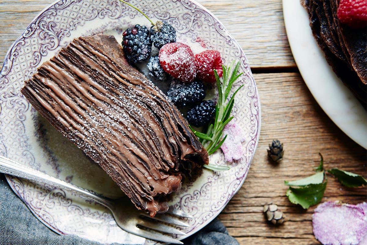 A slice of chocolate crepe cake on a plate, dusted with confectioners' sugar
