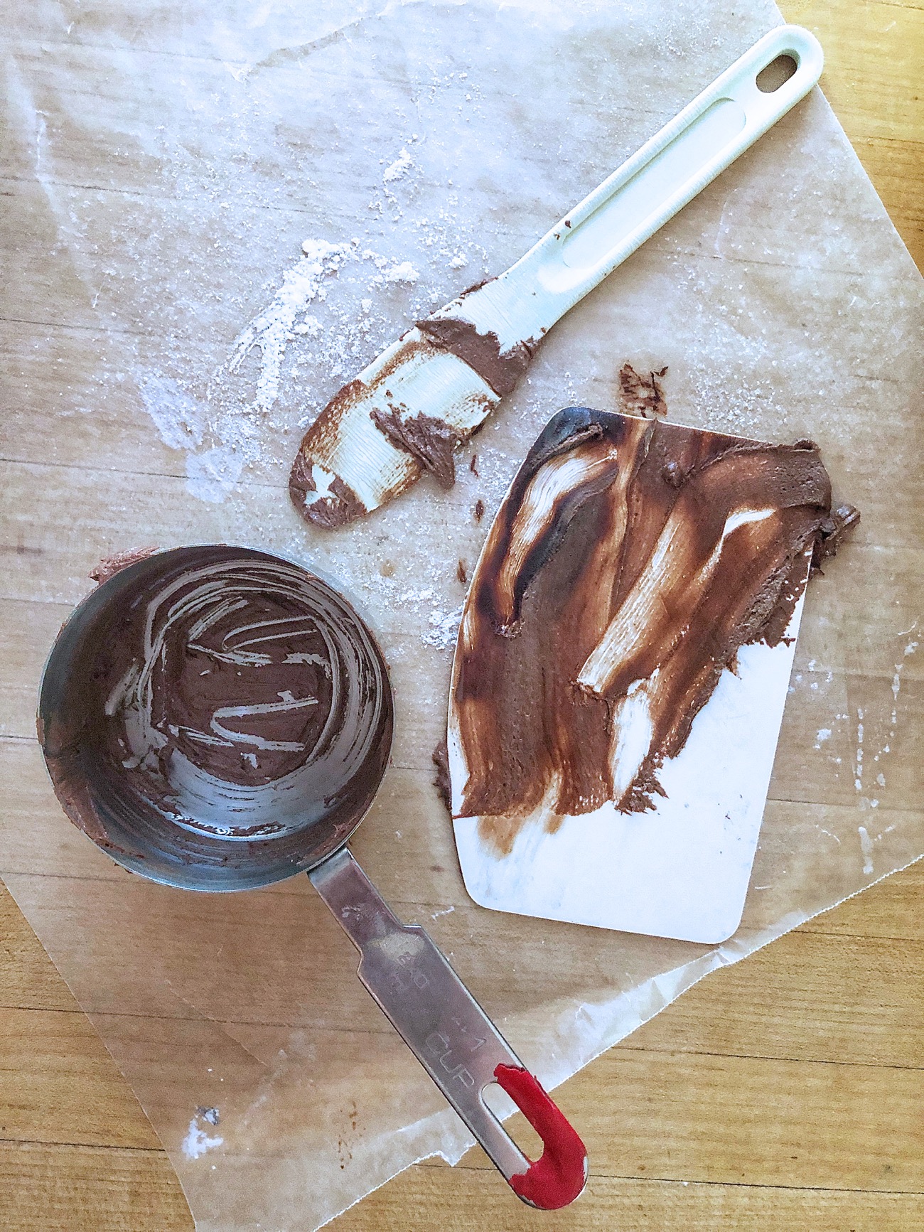 Measuring cup, spatula, and bowl scraper, all covered with chocolate frosting, set on a piece of waxed paper.