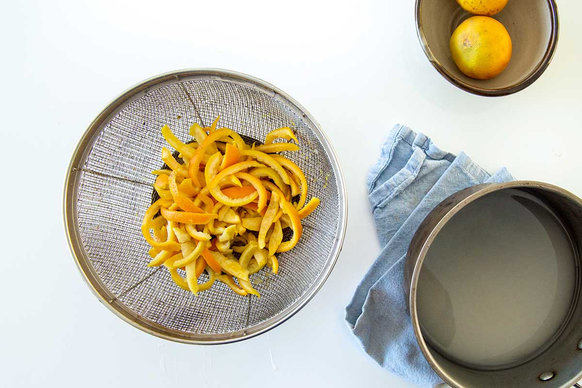 Blanched orange peels in a colander, with a saucepan with sugar and water