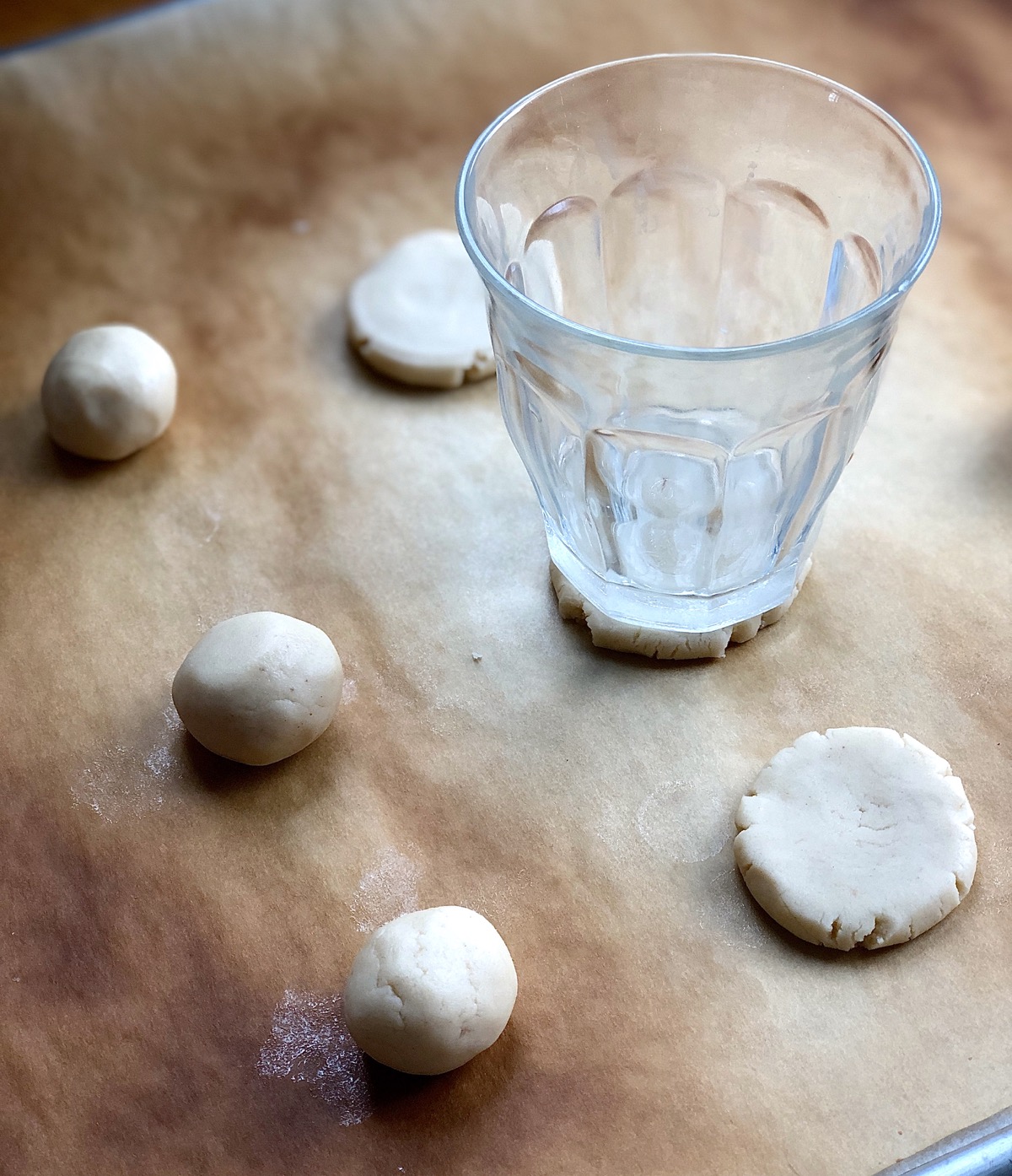 Balls os shortbread dough on a baking sheet, one ball being flattened with the bottom of a drinking glass.
