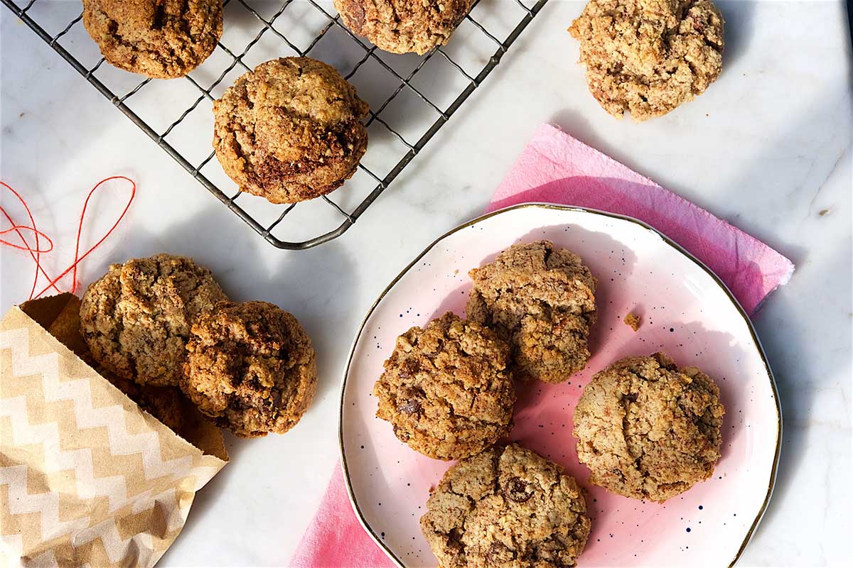 A batch of chocolate chip and chunk cookies on a kitchen table with small paper bags for gifting